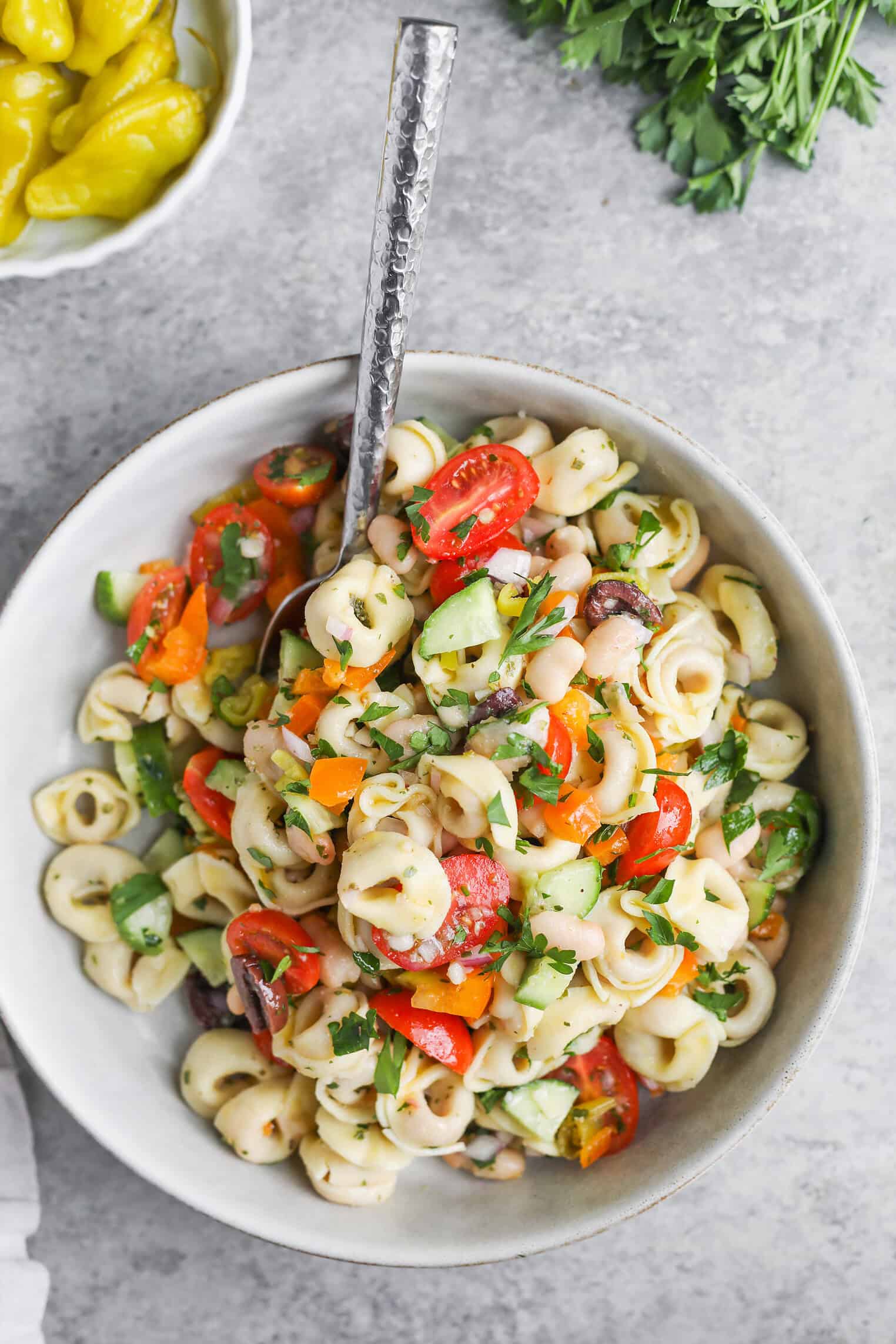 Tortellini Pasta Salad 1 A Bowl Of Tortellini Pasta Salad With Cherry Tomatoes, Cucumber, Bell Pepper, Olives, And Parsley Is Served With A Fork. Fresh Parsley And A Bowl Of Pepperoncini Accompany This Vibrant Tortellini Pasta Salad On The Side.