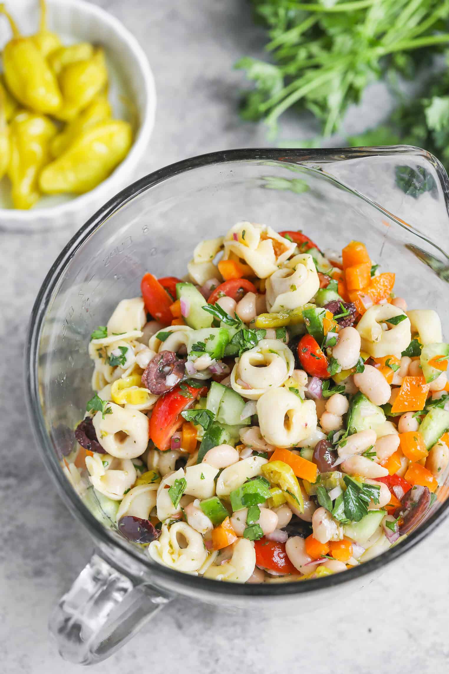 Tortellini Pasta Salad 2 A Glass Bowl Filled With Vibrant Tortellini Pasta Salad, Featuring Chopped Vegetables, Beans, And Herbs. Fresh Parsley And A Small Bowl Of Yellow Peppers Sit In The Background On A Gray Surface.