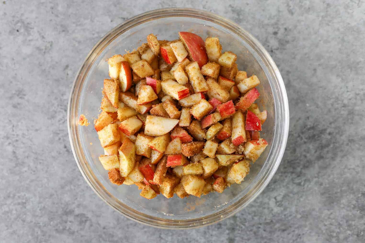 Vegan Apple Cake 5 A Glass Bowl Filled With Diced Apples Coated In Cinnamon And Sugar, Reminiscent Of The Flavors In A Cinnamon Spice Apple Cake, Sits On A Gray Textured Surface.