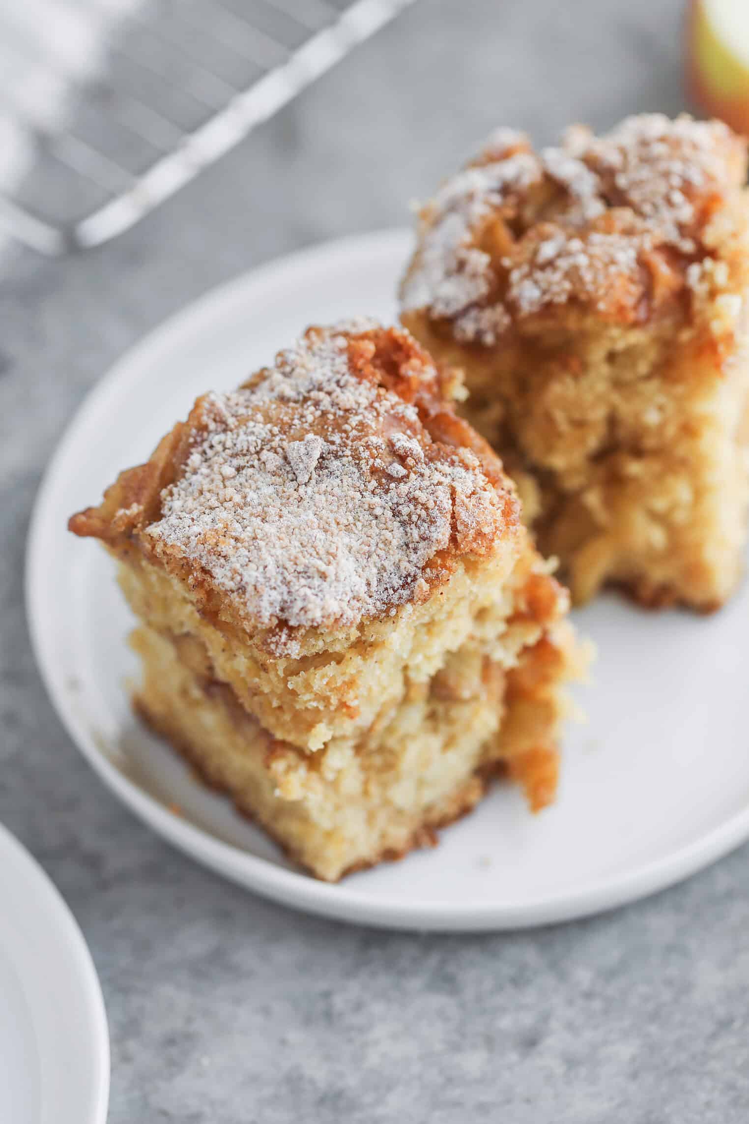 Vegan Apple Cake 9 Two Pieces Of Moist, Crumbly Cinnamon Spice Apple Cake Topped With Powdered Sugar Sit On A White Plate, With A Cooling Rack And Part Of Another Plate Visible In The Background.