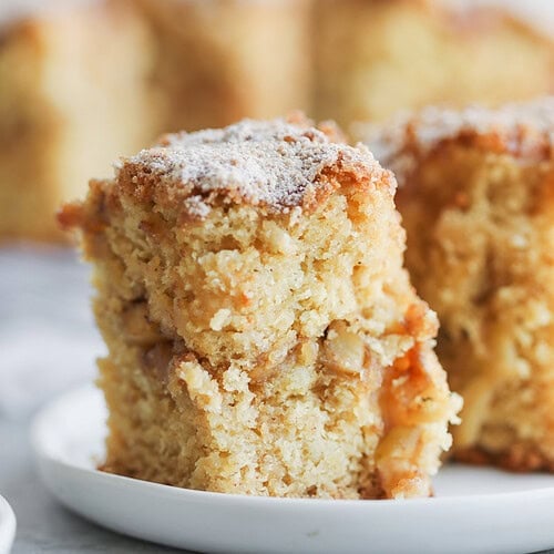 Vegan Apple Cake 12 A Close-Up Of A Thick Slice Of Cinnamon Spice Apple Cake With A Crumbly Topping, Sitting On A White Plate. The Cake Looks Moist With Visible Apple Chunks, And Another Piece Is Blurred In The Background.