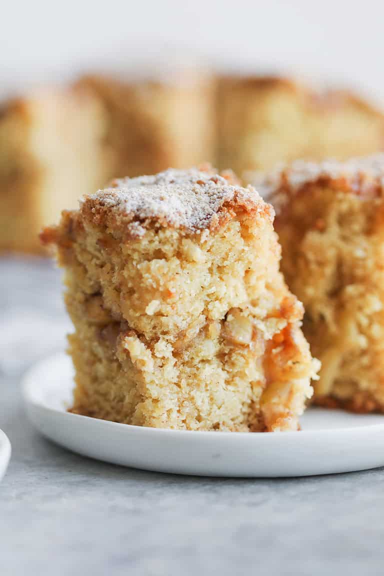 A Close-Up Of A Thick Slice Of Cinnamon Spice Apple Cake With A Crumbly Topping, Sitting On A White Plate. The Cake Looks Moist With Visible Apple Chunks, And Another Piece Is Blurred In The Background.