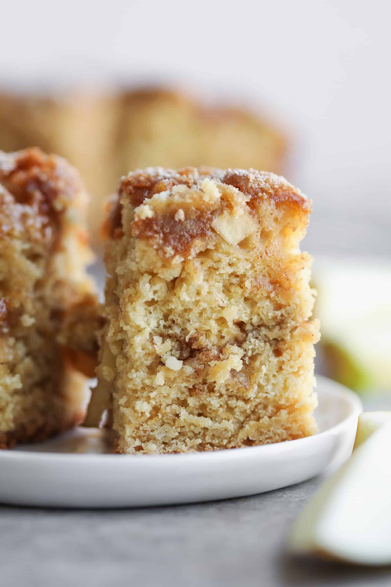Vegan Apple Cake 10 A Close-Up Of A Thick, Moist Slice Of Cinnamon Spice Apple Cake With A Crumb Topping Sits On A White Plate, While More Cake Pieces And A Green Utensil Are Blurred In The Background.