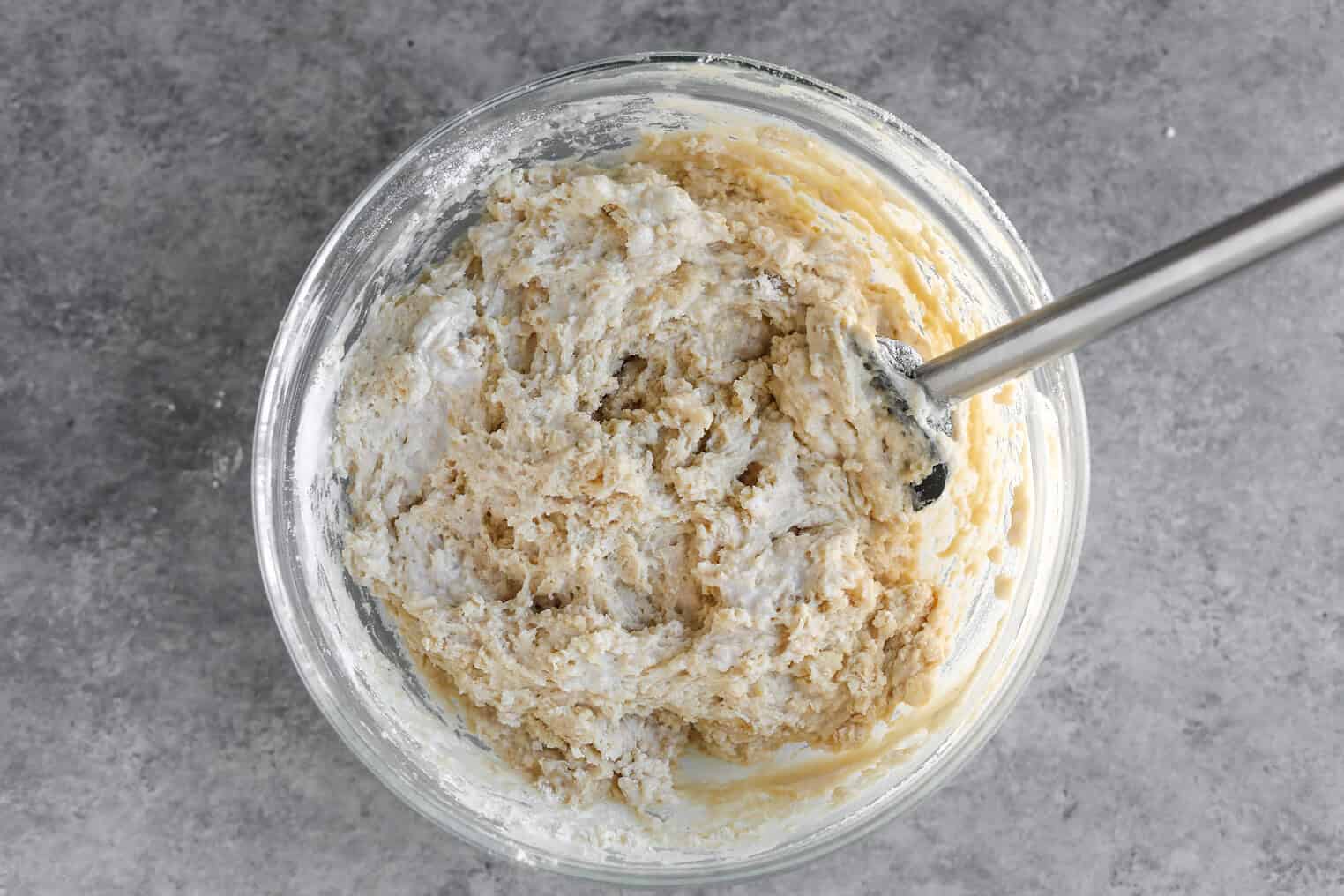 Vegan Apple Cake 6 A Glass Bowl Filled With Thick, Partially Mixed Cinnamon Spice Apple Cake Dough And A Metal Spoon, Sitting On A Gray Countertop.