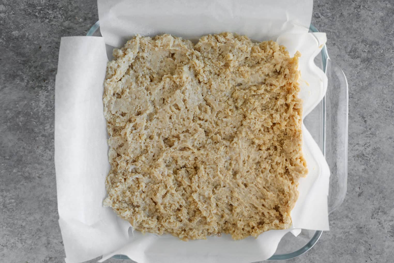 Vegan Apple Cake 7 A Glass Baking Dish Lined With Parchment Paper Holds Unbaked Cinnamon Spice Apple Cake Dough Spread Evenly Inside. The Dish Sits On A Gray Countertop.