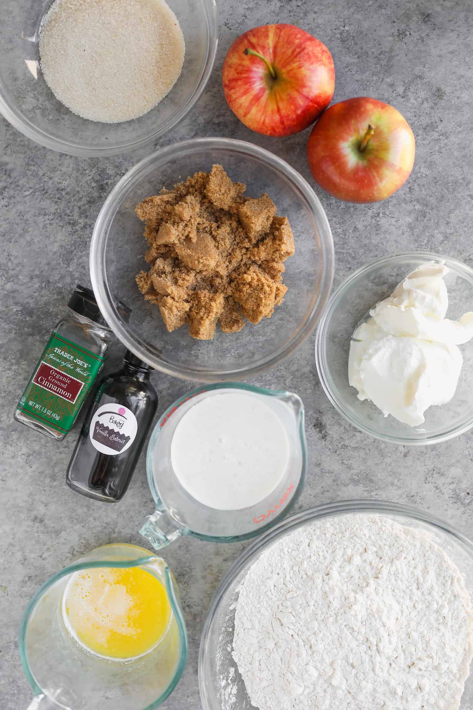 Vegan Apple Cake 2 Ingredients For Vegan Apple Cake. Bowls Of Flour, Brown Sugar, Granulated Sugar, And Sour Cream, Along With Ingredients For Cinnamon Spice Apple Cake—Milk, Melted Butter, Apples, Ground Cinnamon, And Vanilla Extract—Are Arranged On A Gray Countertop.