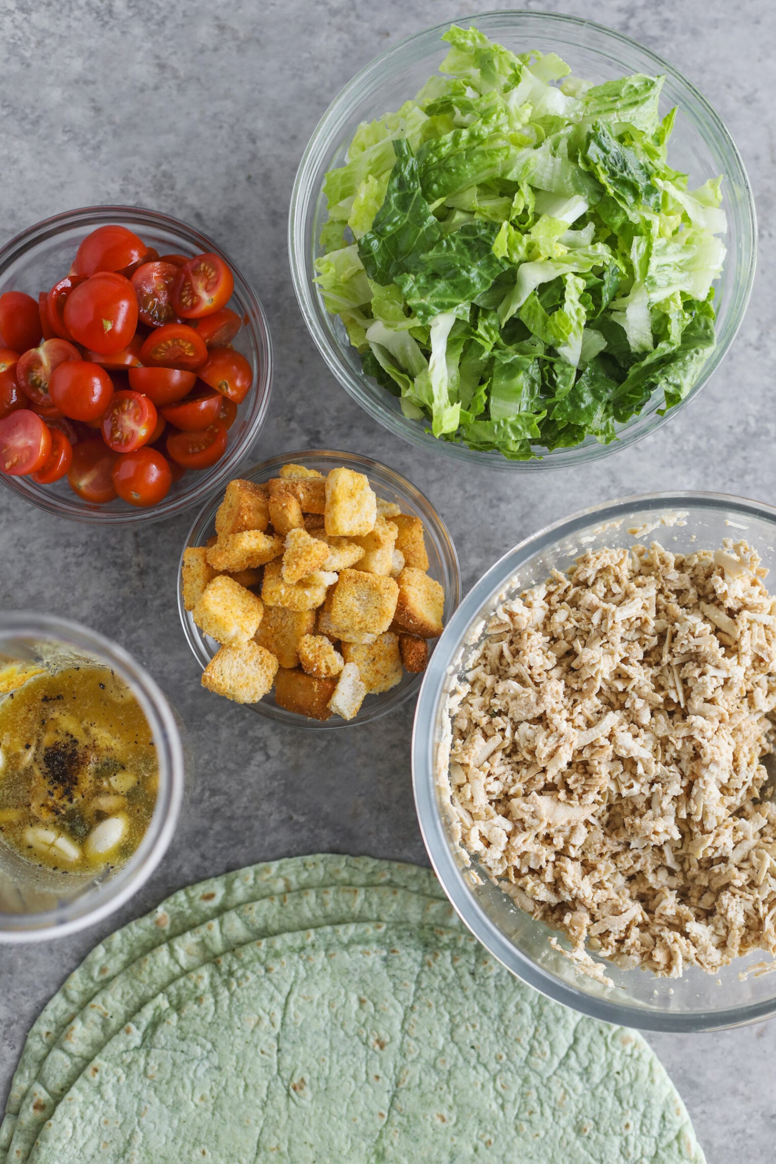 Five Bowls With Salad Ingredients: Chopped Romaine Lettuce, Halved Cherry Tomatoes, Croutons, Vegan Chicken Caesar Strips, And A Mixture Of Dressing. Several Spinach Tortillas Are Laid Out On The Grey Countertop.