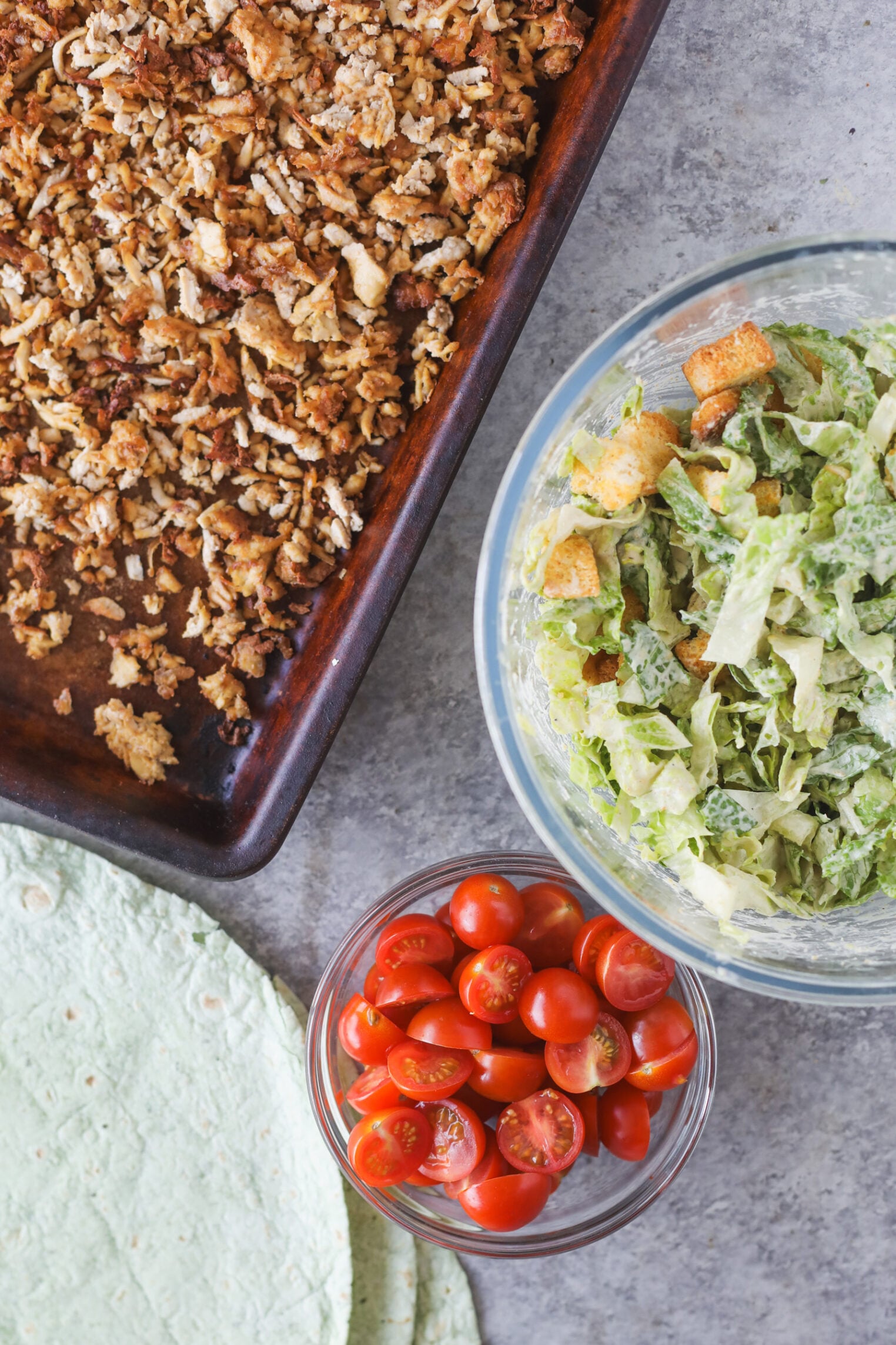 A Baking Tray With Crispy Seasoned Vegan Chicken Caesar Crumbles, A Bowl Of Chopped Romaine Lettuce With Croutons, A Bowl Of Halved Cherry Tomatoes, And A Spinach Tortilla Wrap On A Gray Surface.
