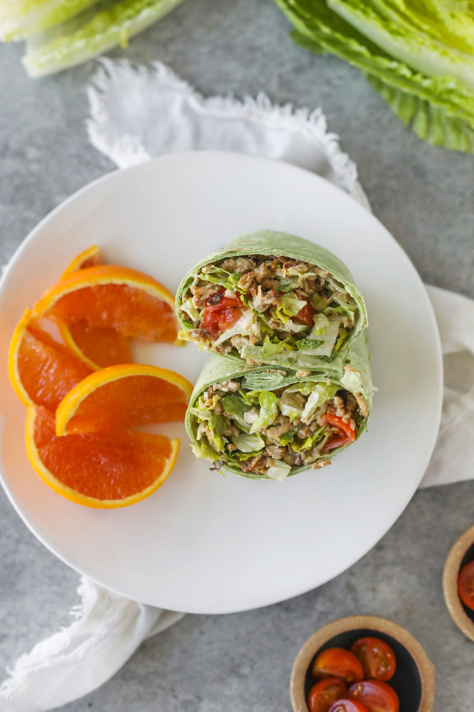 A Spinach Wrap Stuffed With Vegan Chicken Caesar, Lettuce, And Tomatoes Sits Halved On A White Plate, Accompanied By Three Orange Slices. There Are Lettuce Leaves, A Napkin, And Bowls Of Cherry Tomatoes Nearby.