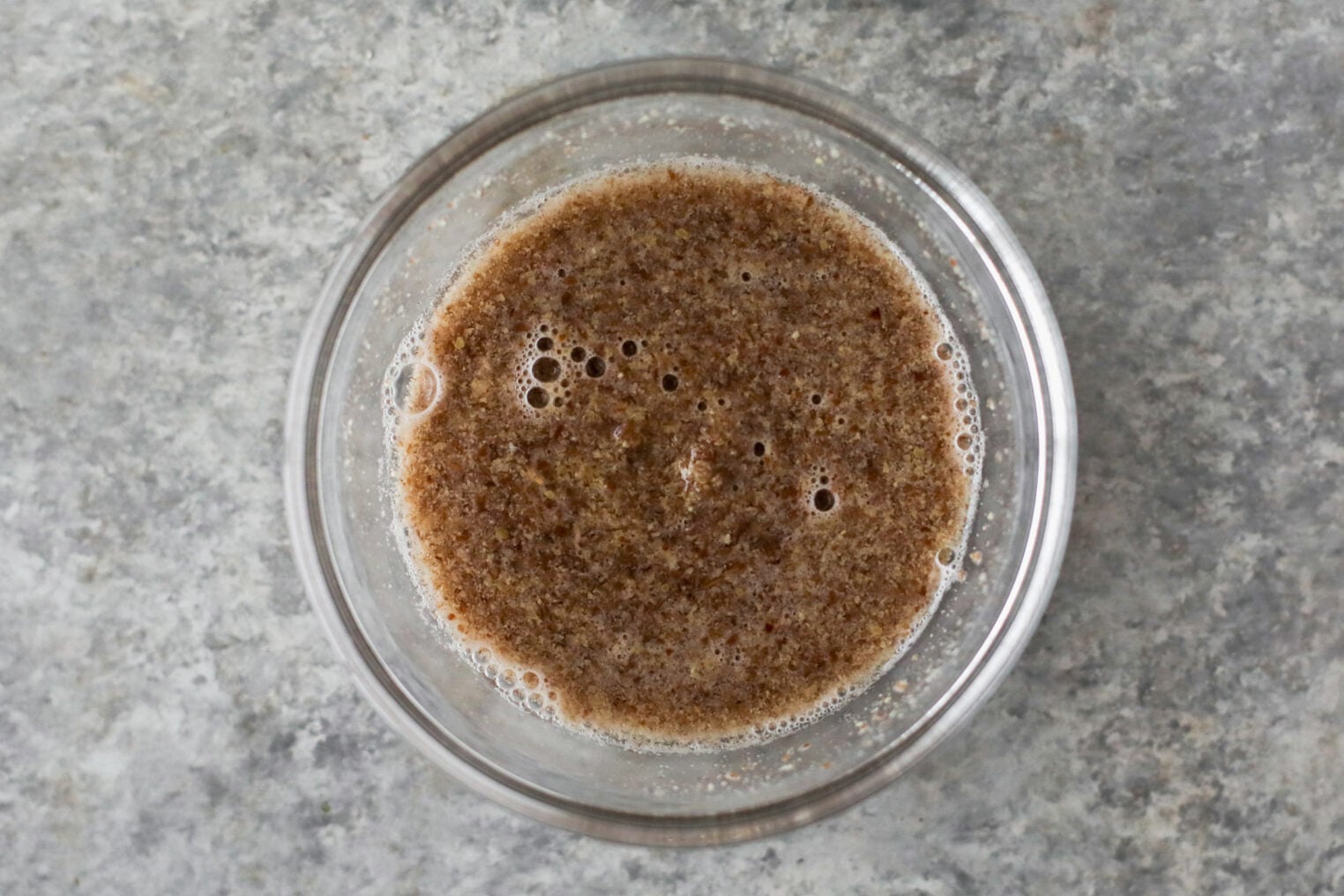 A Glass Bowl Filled With A Brown, Speckled Liquid Mixture—Likely Ground Flaxseed And Water—Sits On A Gray Textured Countertop, Ready To Be Used As An Egg Substitute For The Best Vegan Chocolate Chip Cookies.