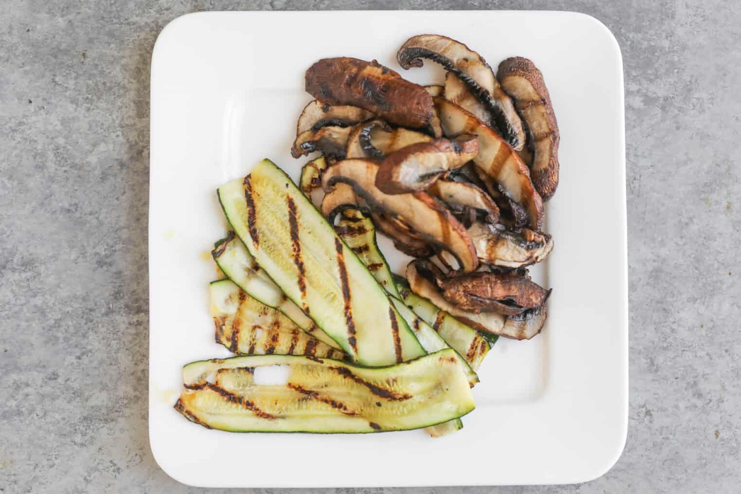 Vegan Italian Panini 6 A Square White Plate With Grilled Slices Of Zucchini And Portobello Mushrooms, Arranged Side By Side As Delicious Vegan Panini Fillings, On A Gray Stone Surface.