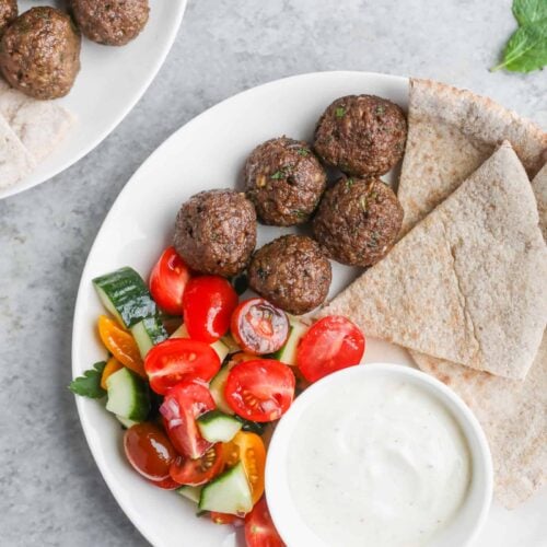 A Plate With Vegan Kofta Meatballs, Sliced Pita Bread, A Colorful Salad Of Cucumbers And Tomatoes, And A Bowl Of Creamy White Sauce. Another Plate With Similar Food Is Partially Visible In The Background. Mint Leaves Are Scattered On A Gray Surface.