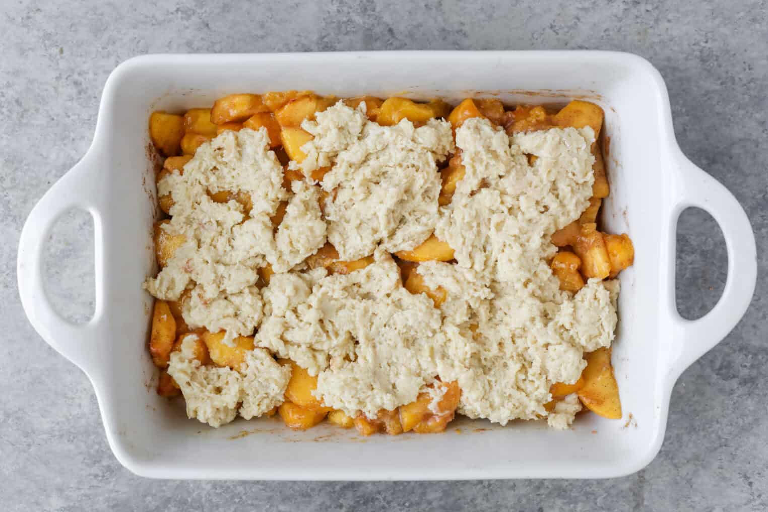 A White Baking Dish Filled With Vegan Peach Cobbler, Featuring Sliced Peaches On The Bottom And Rough Spoonfuls Of Biscuit Dough Scattered On Top, Ready To Be Baked.