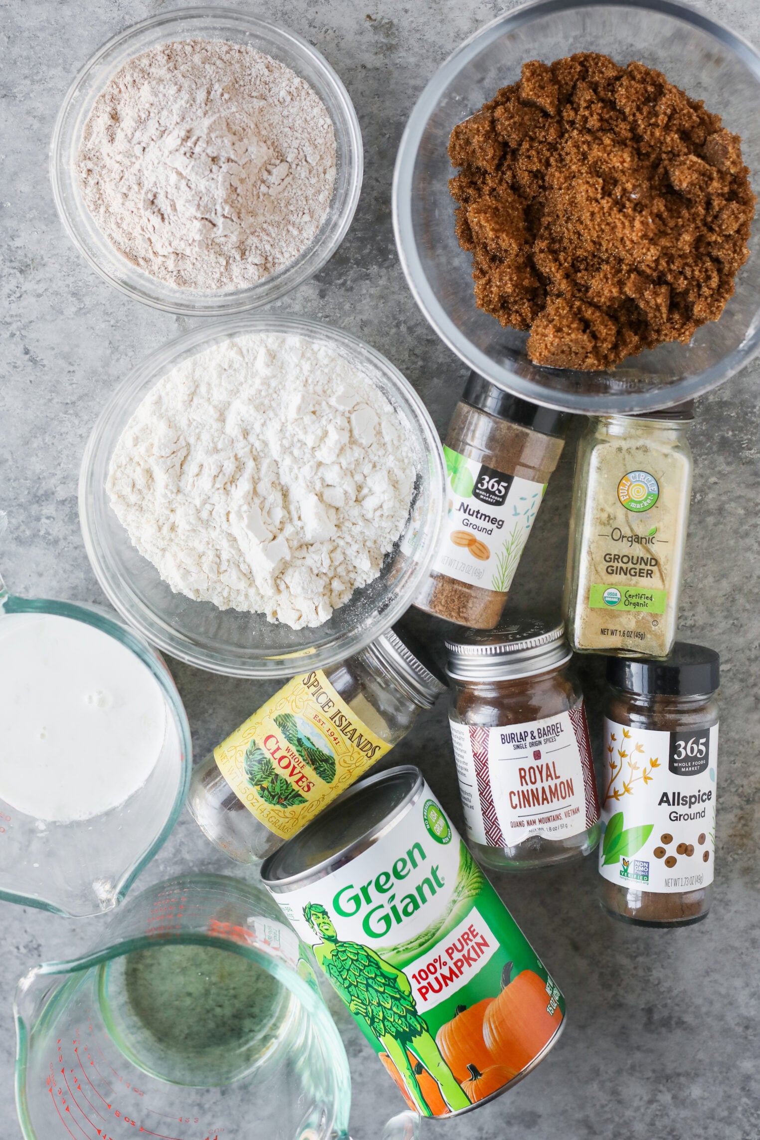 Ingredients For A Vegan Pumpkin Bread Recipe Arranged On A Gray Surface, Including Bowls Of Flour And Brown Sugar, A Can Of Pumpkin Puree, Milk, And Jars Of Nutmeg, Ginger, Cloves, Cinnamon, And Allspice.