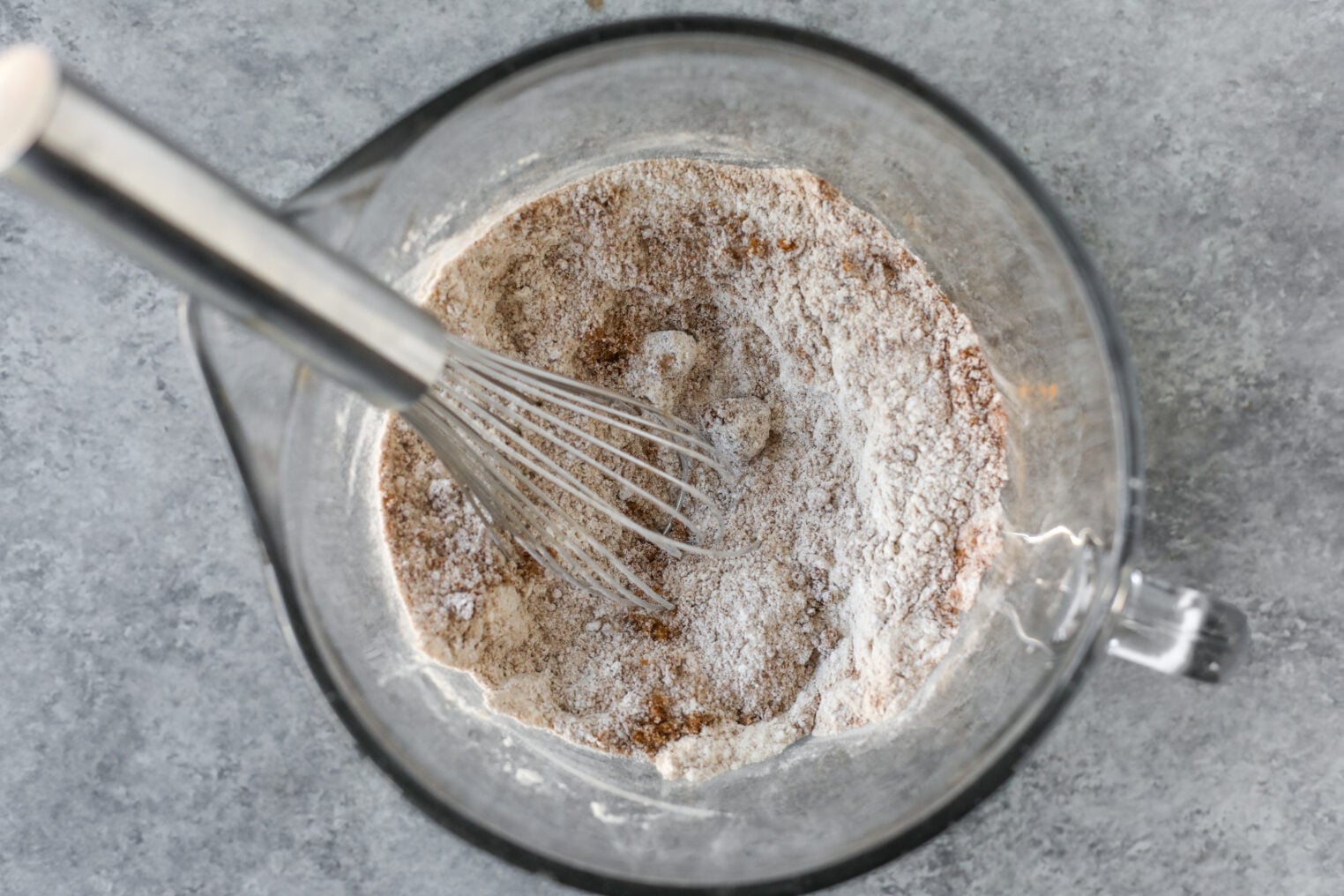 A Whisk Sits In A Glass Mixing Bowl Containing Flour And Other Dry Ingredients For Vegan Pumpkin Bread.