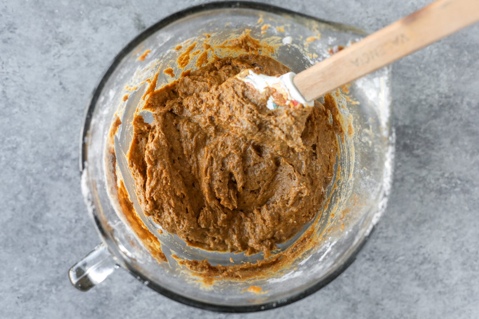 A Silicone Spatula Mixing Thick Brown Vegan Pumpkin Bread Dough In A Glass Bowl.