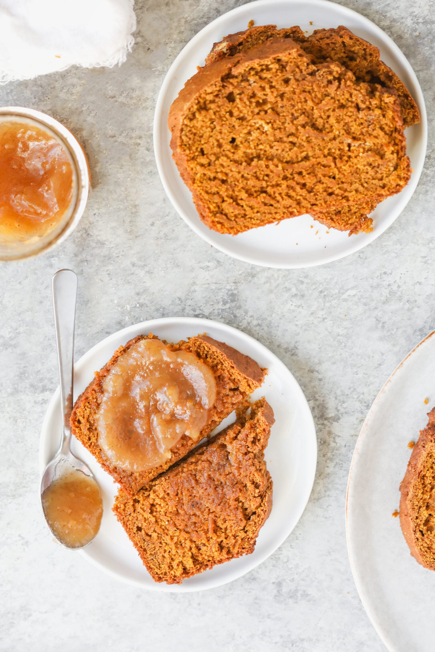 Slices Of Vegan Pumpkin Bread On Small Plates, With One Slice Topped With Apple Butter Next To A Spoon Of Apple Butter.