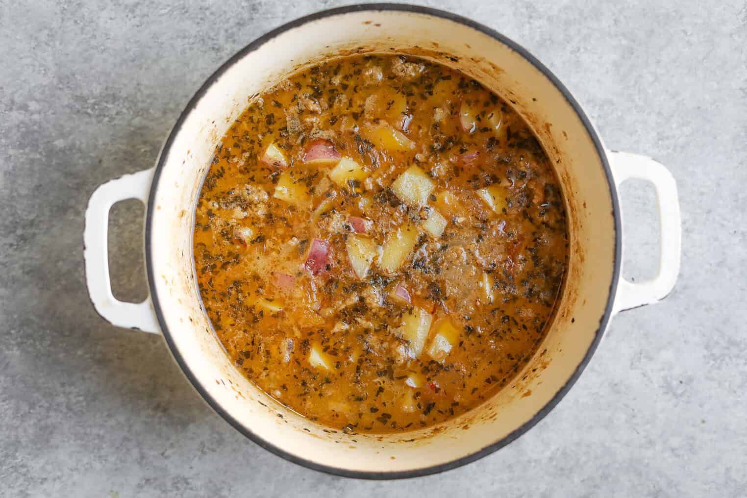 Potato, Spinach and Sausage Soup 5 A White Pot Filled With Hearty Spinach Sausage Soup, Featuring Diced Potatoes, Ground Meat, Herbs, And A Golden Broth, Sits On A Gray Textured Surface.
