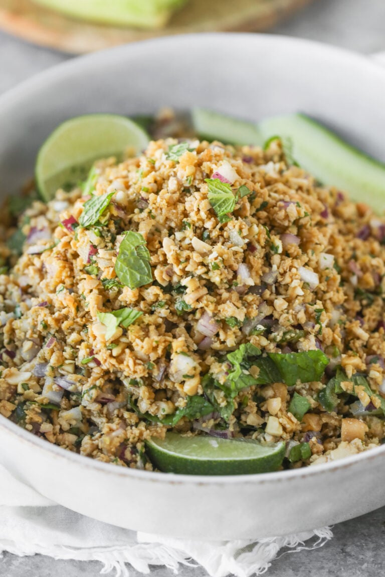 A Close-Up Of A Bowl Filled With A Chopped Salad Made From Cauliflower, Herbs, Onions, And Other Vegetables—Perfect For Vegan Thai Larb Lettuce Wraps—Garnished With Fresh Mint And Lime Wedges.