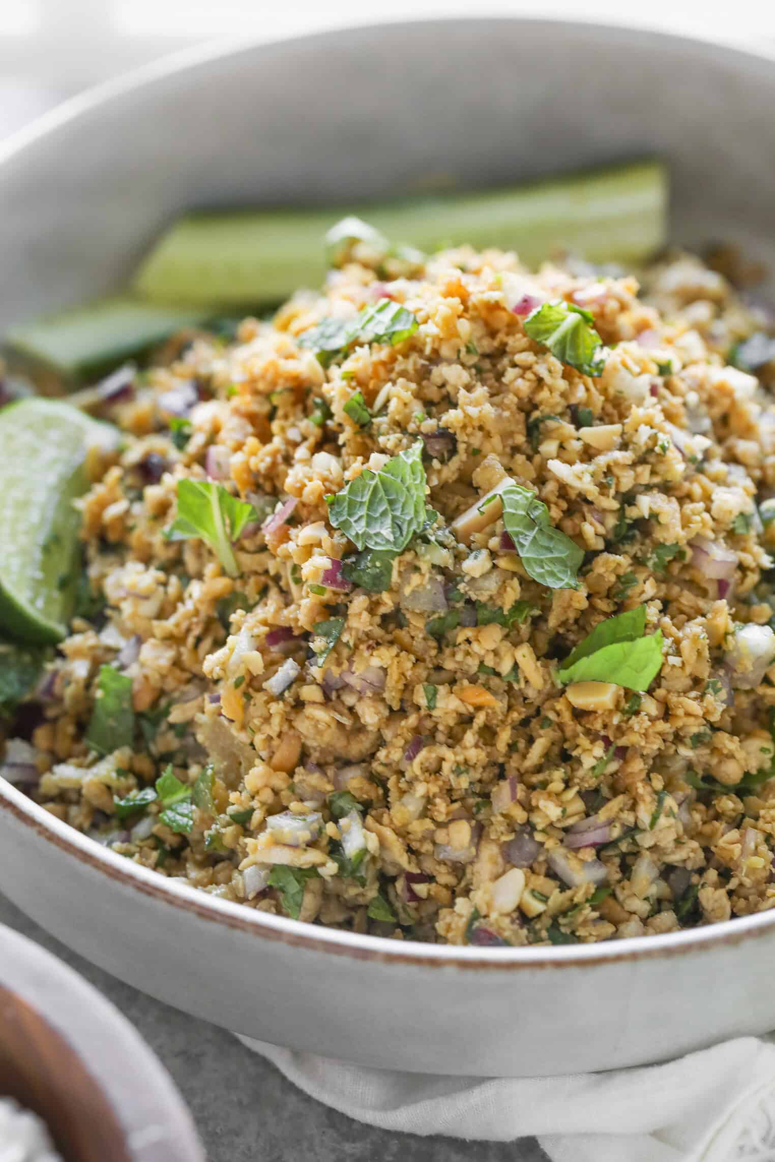 Vegan Thai Larb Lettuce Wraps 3 A Bowl Of Crispy Rice Salad Inspired By Vegan Thai Larb, Mixed With Herbs, Chopped Onions, And Mint Leaves, Garnished With Fresh Mint And Lime Wedges, With Cucumber Slices On The Side.