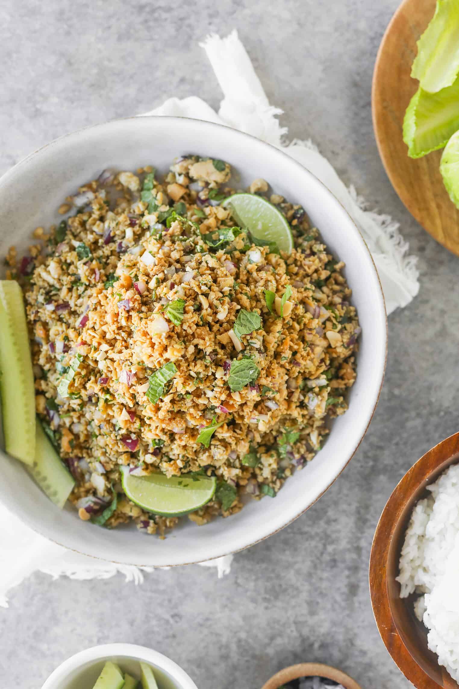 Vegan Thai Larb Lettuce Wraps 2 A Bowl Of Vegan Thai Larb Lettuce Wraps, A Minced Plant-Based Salad Garnished With Mint And Lime Wedges, Sits On A Gray Surface With Sliced Cucumber On The Side. Other Dishes And Lettuce Leaves Are Partially Visible Nearby.