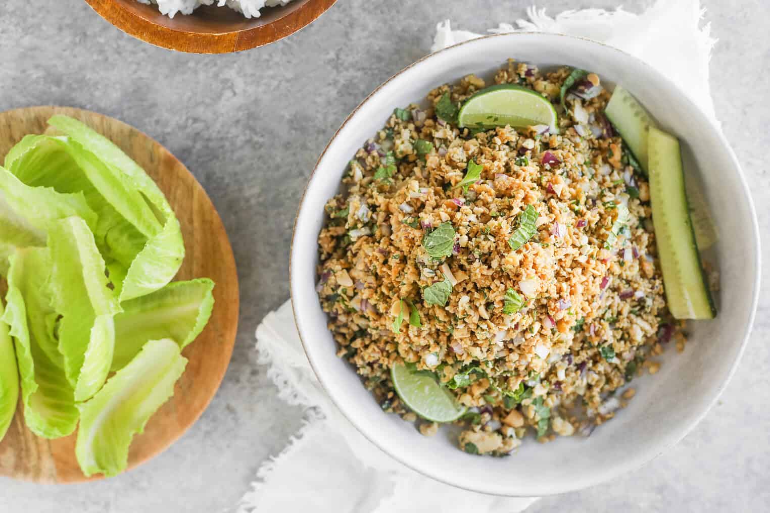 Vegan Thai Larb Lettuce Wraps 6 A Bowl Of Vegan Thai Larb Cauliflower Rice Salad With Herbs, Red Onion, Lime Wedges, And Cucumber Slices Sits On A White Cloth. Next To It Is A Wooden Plate Filled With Fresh Romaine Lettuce Leaves.