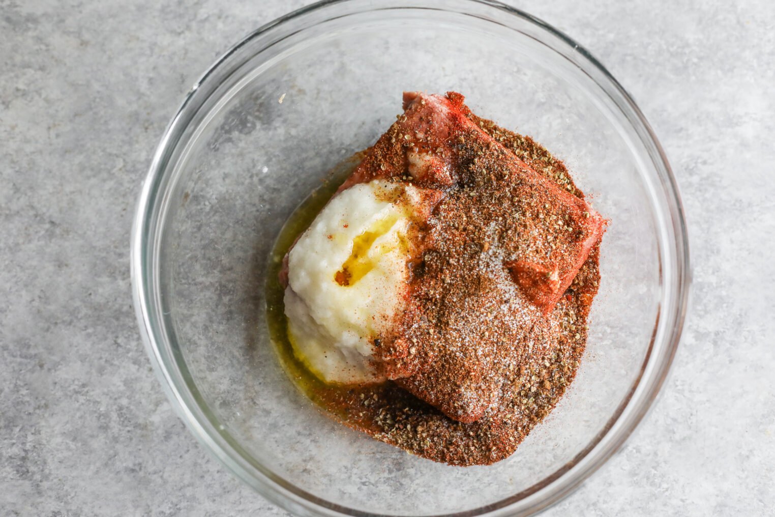 A Glass Bowl Containing Raw Ground Vegan Doner Kebab Mix, Finely Chopped Onions, Oil, And A Generous Amount Of Mixed Spices, Set On A Light Gray Surface.