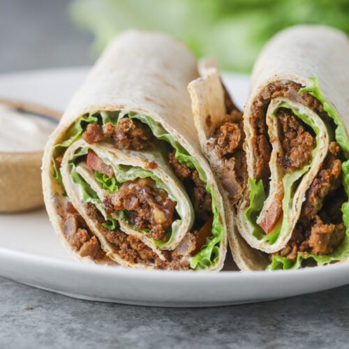 Two Halves Of A Turkish Doner Wrap Filled With Seasoned Ground Meat, Lettuce, And Tomato Are Placed On A White Plate, With A Blurred Background Of Lettuce And A Small Bowl Of Sauce.