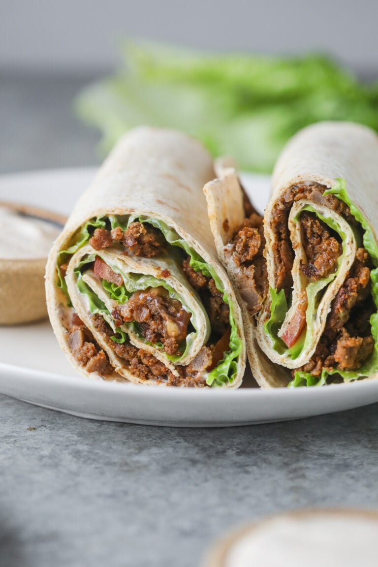Two Halves Of A Turkish Doner Wrap Filled With Seasoned Ground Meat, Lettuce, And Tomato Are Placed On A White Plate, With A Blurred Background Of Lettuce And A Small Bowl Of Sauce.