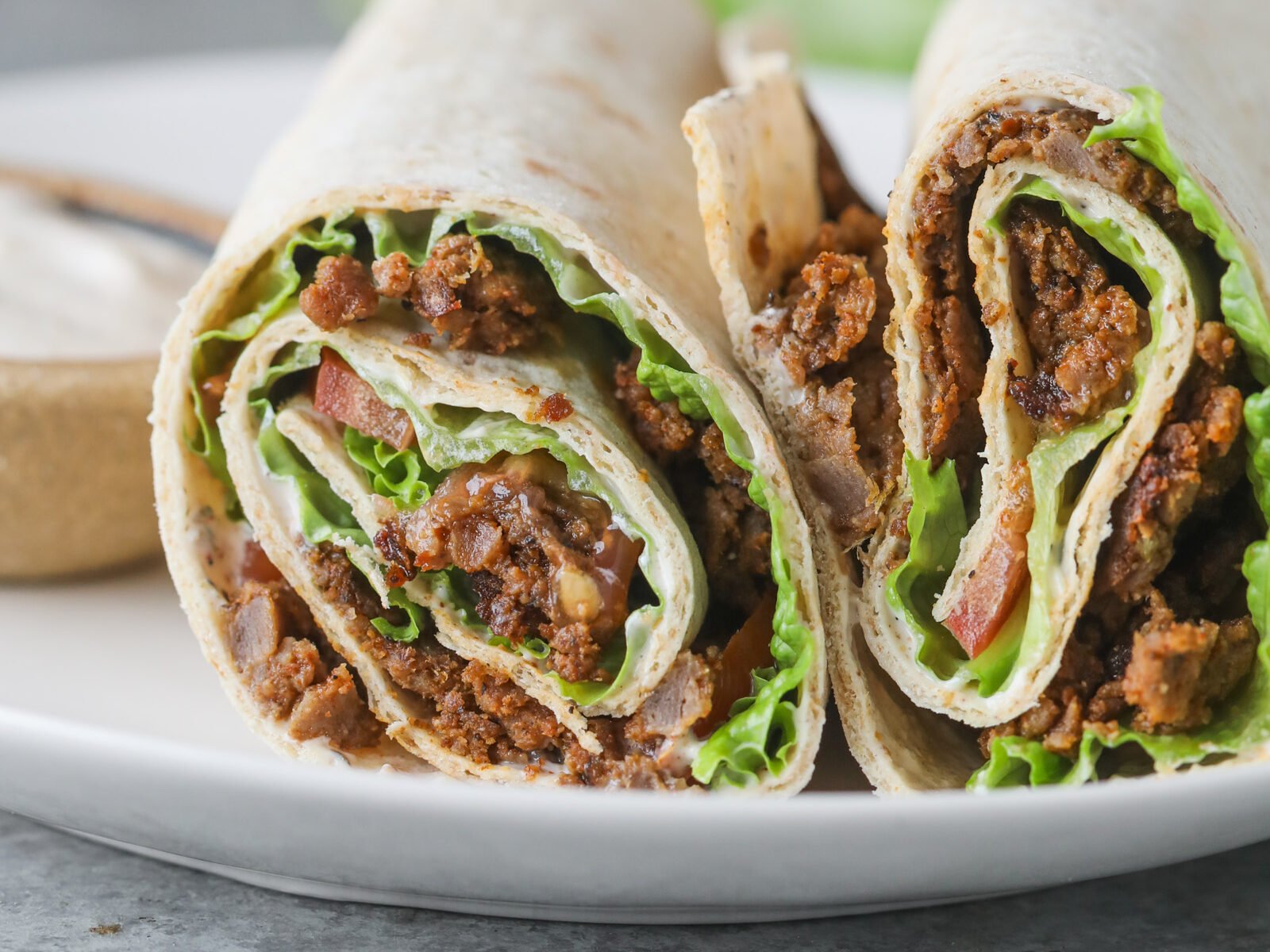 Two Halves Of A Turkish Doner Wrap Filled With Seasoned Ground Meat, Lettuce, And Tomato Are Placed On A White Plate, With A Blurred Background Of Lettuce And A Small Bowl Of Sauce.