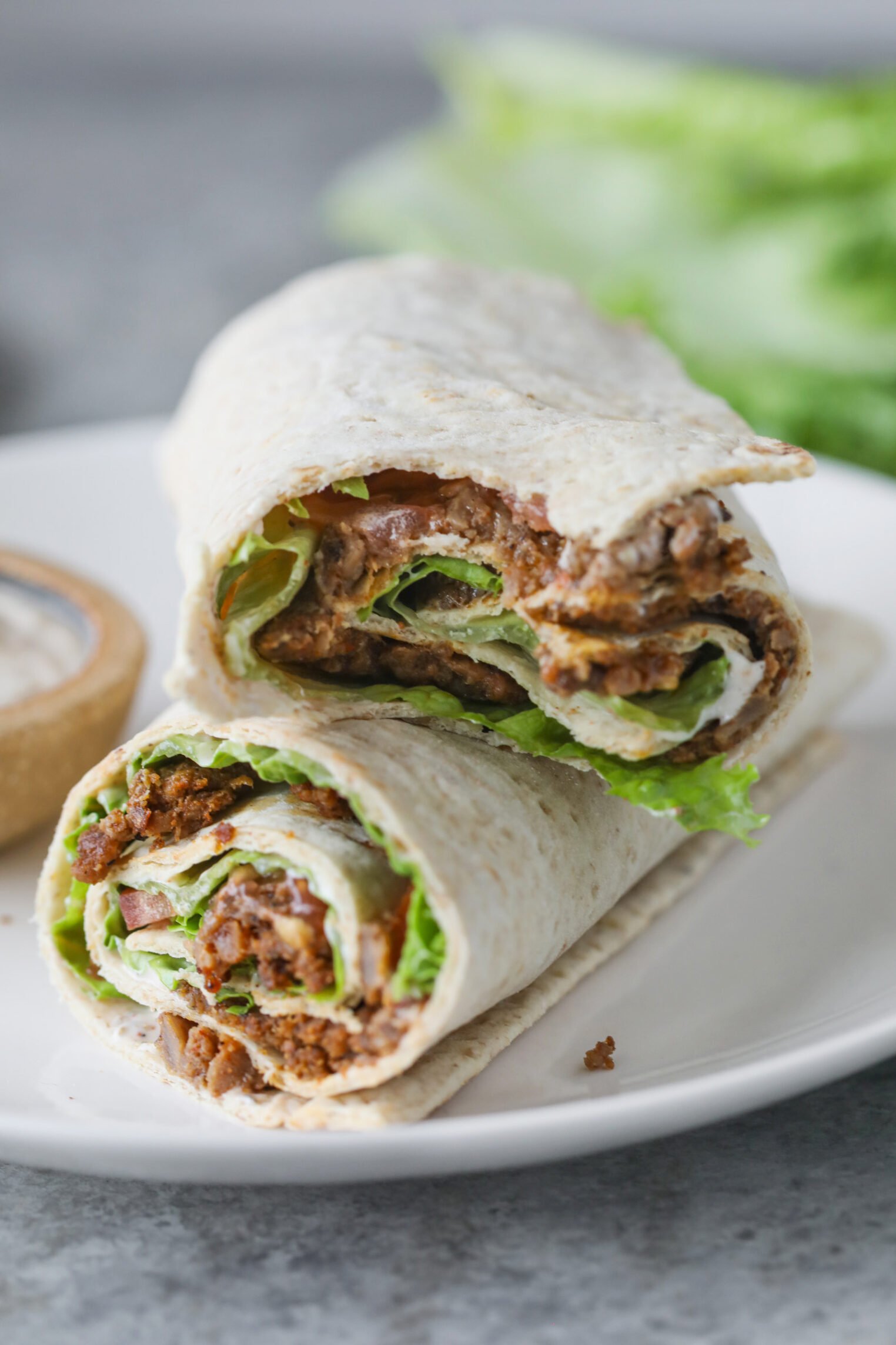 A Close-Up Of A Vegan Doner Kebab Wrap Sandwich Cut In Half, Filled With Seasoned Plant-Based Meat, Lettuce, And Sauce, Served On A White Plate With A Small Bowl Of Dipping Sauce In The Background.