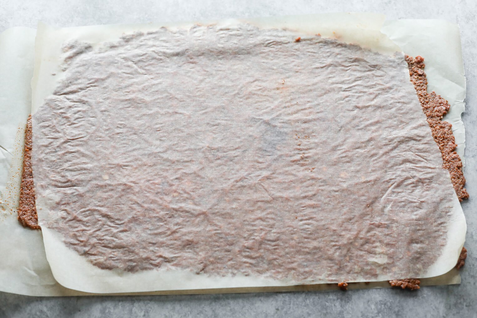 A Baking Sheet Covered With A Thin Layer Of Vegan Doner Kebab Mixture, Partially Covered By Parchment Paper, Is Shown On A Gray Surface. Some Of The Savory Mixture Is Visible Peeking Out From The Edges Of The Paper.