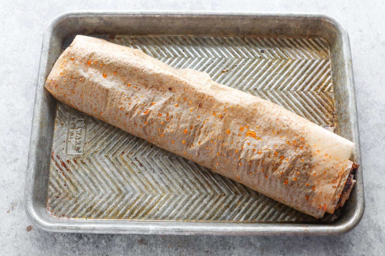 A Large Rolled Pastry, Reminiscent Of A Vegan Doner Kebab, Is Wrapped In Parchment Paper And Rests On A Metal Baking Sheet, With Small Spots Of Oil Visible On The Surface.