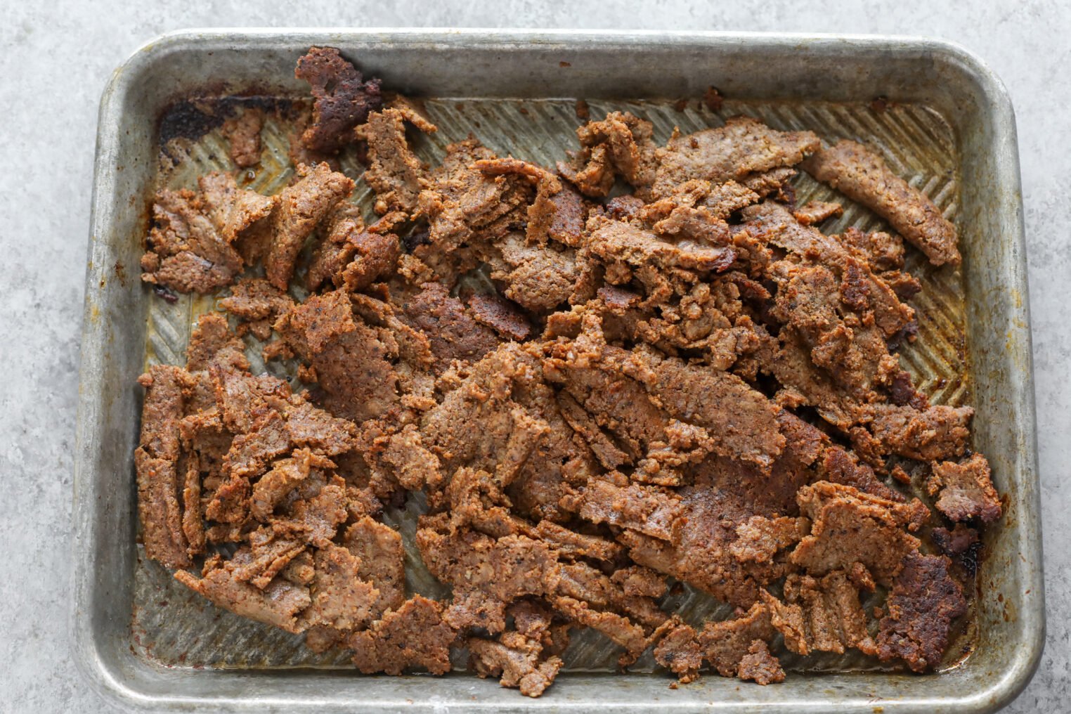 Thinly Sliced, Cooked Vegan Doner Kebab Seitan Pieces Spread Out On A Metal Baking Tray With A Textured Surface, Set On A Light Gray Countertop. The Seitan Appears Browned And Slightly Crispy.