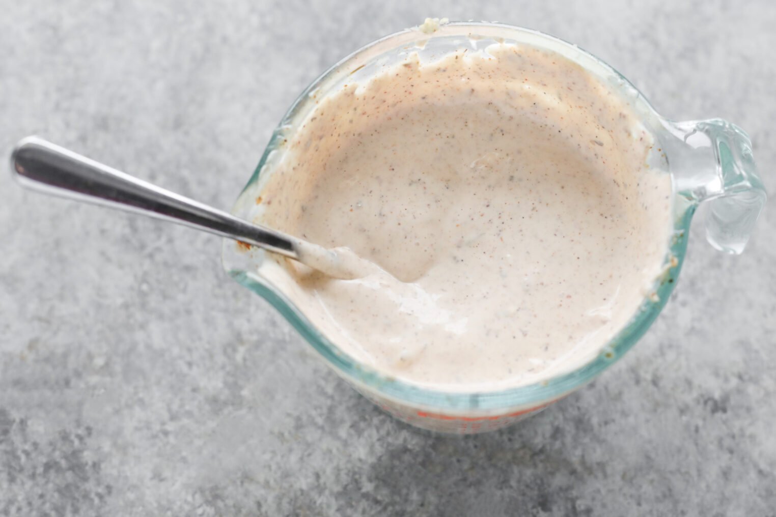 A Glass Measuring Cup Filled With A Creamy, Speckled Vegan Doner Kebab Sauce Or Dressing, With A Metal Spoon Resting In It, Placed On A Textured Gray Surface.