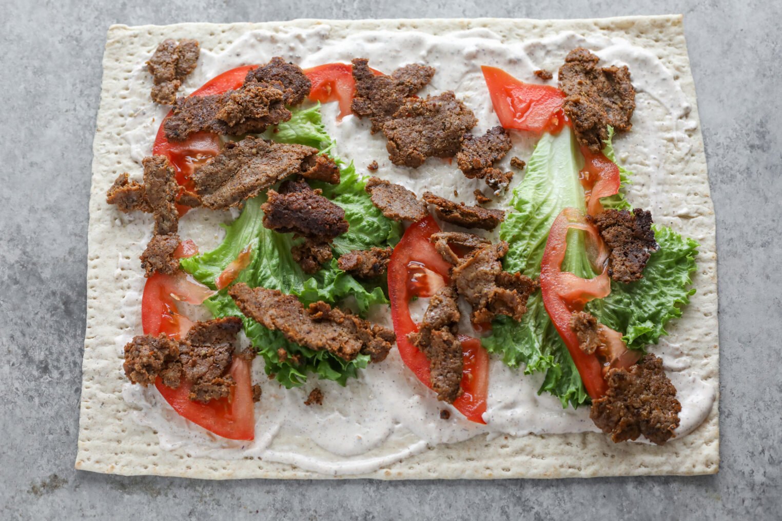 A Rectangular Vegan Doner Kebab Flatbread Topped With A Spread Of White Sauce, Leafy Green Lettuce, Sliced Tomatoes, And Pieces Of Seasoned Cooked Meat On A Gray Surface.