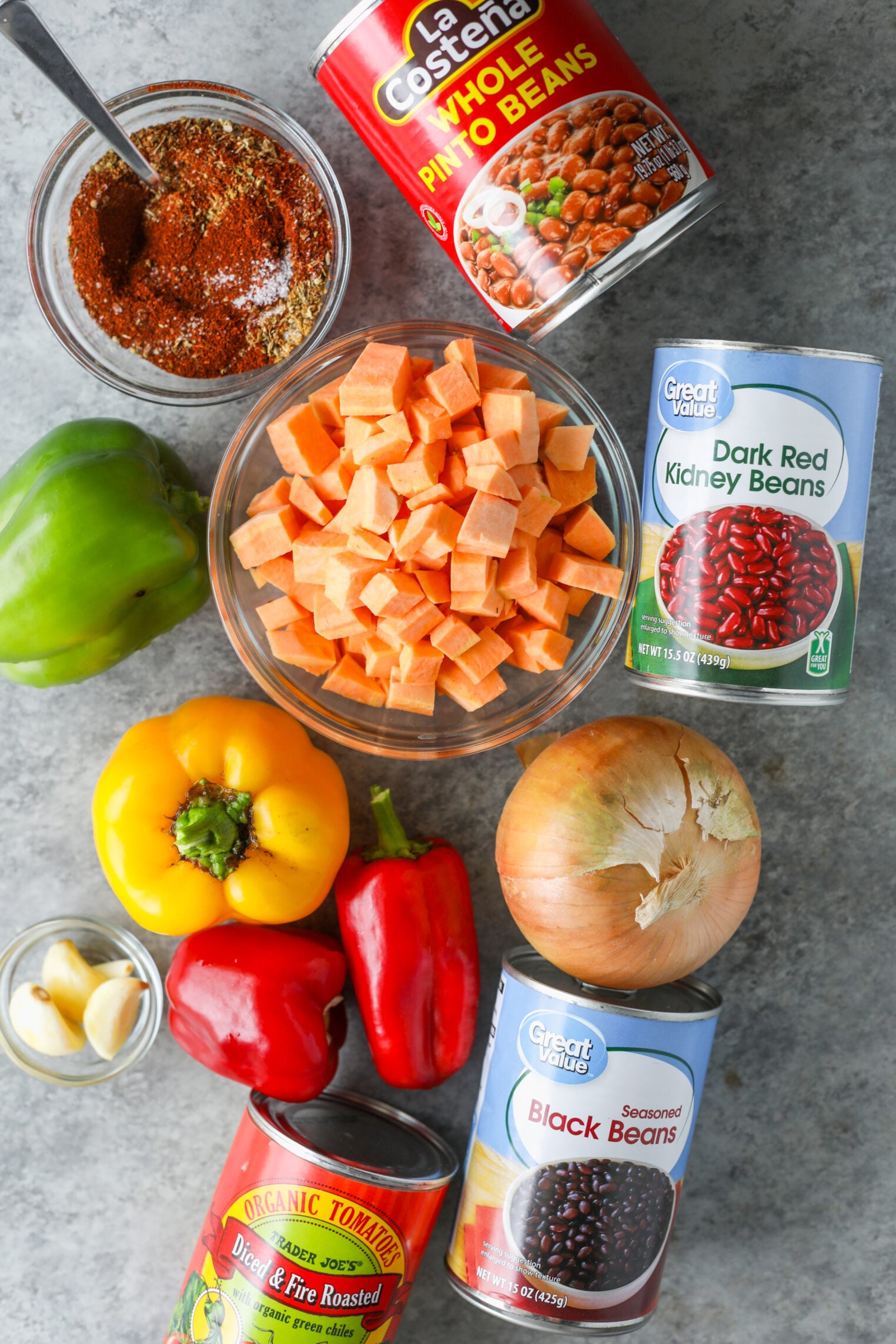 Ingredients For Vegetarian Chili Including Cubed Sweet Potatoes, Canned Pinto, Kidney, And Black Beans, Bell Peppers, An Onion, Garlic, And A Bowl Of Dry Spices.