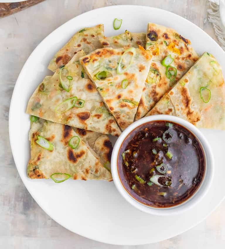 A White Plate Features Scallion Pancakes, Golden Brown With Vibrant Green Bits, Arranged In A Fan Shape. These Healthy Appetizers Are Served Alongside A Small Bowl Of Dark, Herb-Infused Dipping Sauce, Creating An Inviting Presentation.