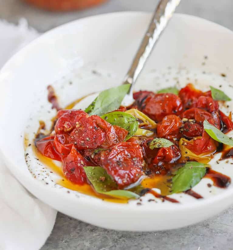 A White Plate Holds A Vibrant Mixture Of Roasted Cherry Tomatoes, Fresh Basil Leaves, And A Drizzle Of Balsamic Glaze, Creating A Delightful Healthy Appetizer. A Spoon Rests On The Plate Amid A Light Pepper Sprinkle, With A White Cloth Partially Visible On The Side.