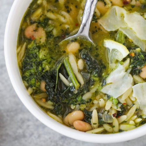 A Bowl Of Bean Orzo Soup With White Beans, Orzo Pasta, Kale, And Shaved Cheese On Top, With A Spoon In The Bowl. A Small Dish Of Red Pepper Flakes Sits Nearby On A Gray Surface.