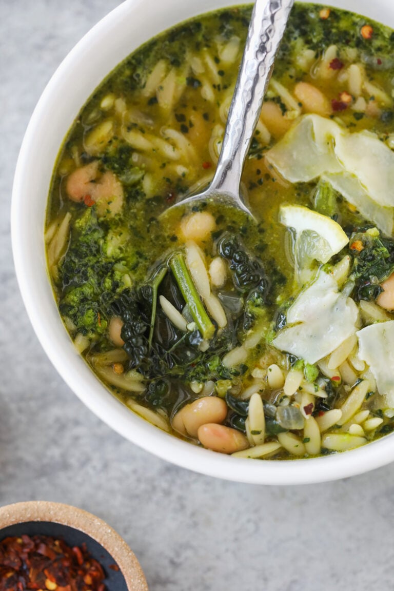 A Bowl Of Bean Orzo Soup With White Beans, Orzo Pasta, Kale, And Shaved Cheese On Top, With A Spoon In The Bowl. A Small Dish Of Red Pepper Flakes Sits Nearby On A Gray Surface.