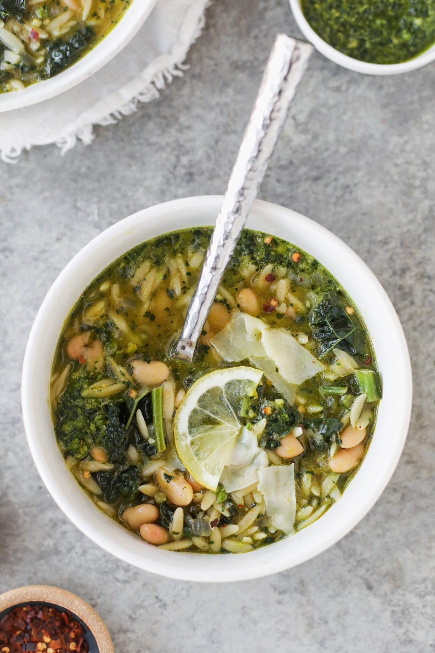 A White Bowl Of Green Bean Orzo Soup With White Beans, Kale, And Spinach, Garnished With Lemon Slices And Shaved Cheese, Sits On A Grey Surface With A Silver Spoon Inside.
