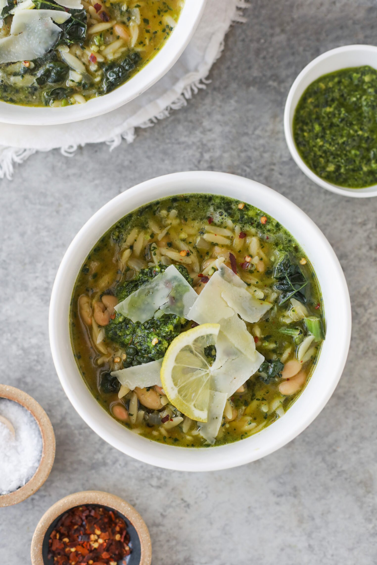 A Bowl Of Green Bean Orzo Soup With Pasta, Beans, Kale, Lemon Slice, And Parmesan Shavings, Garnished With Pesto. Small Bowls Of Salt, Red Pepper Flakes, And Extra Pesto Sit Nearby On A Gray Surface.