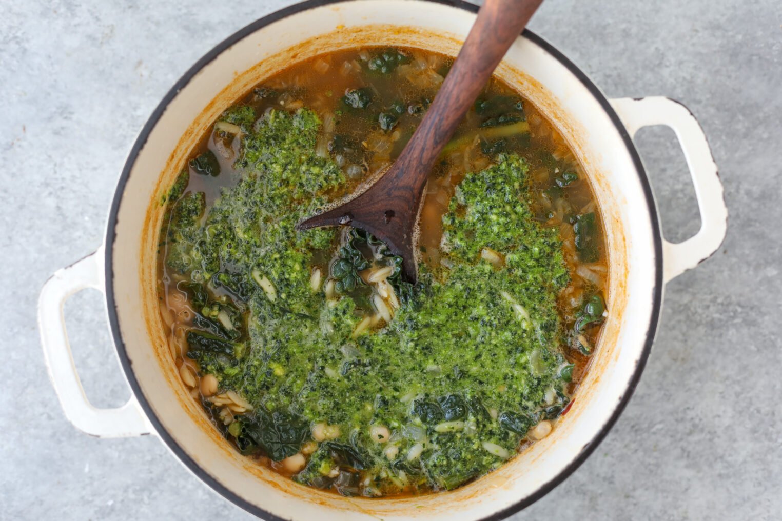 A White Pot Filled With Bean Orzo Soup—Chopped Greens, White Beans, And Orzo Pasta—Topped With A Green Pesto Swirl. A Wooden Spoon Rests Inside The Pot On A Gray Surface.