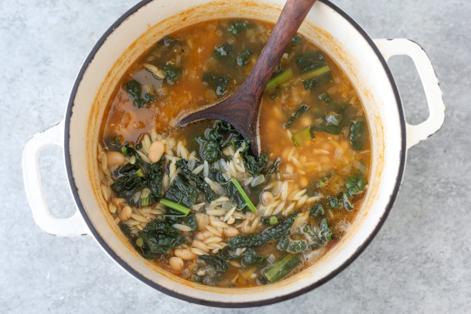 A Pot Of Bean Orzo Soup With Kale, White Beans, Orzo Pasta, And Broth Is Being Stirred With A Wooden Spoon On A Gray Surface.