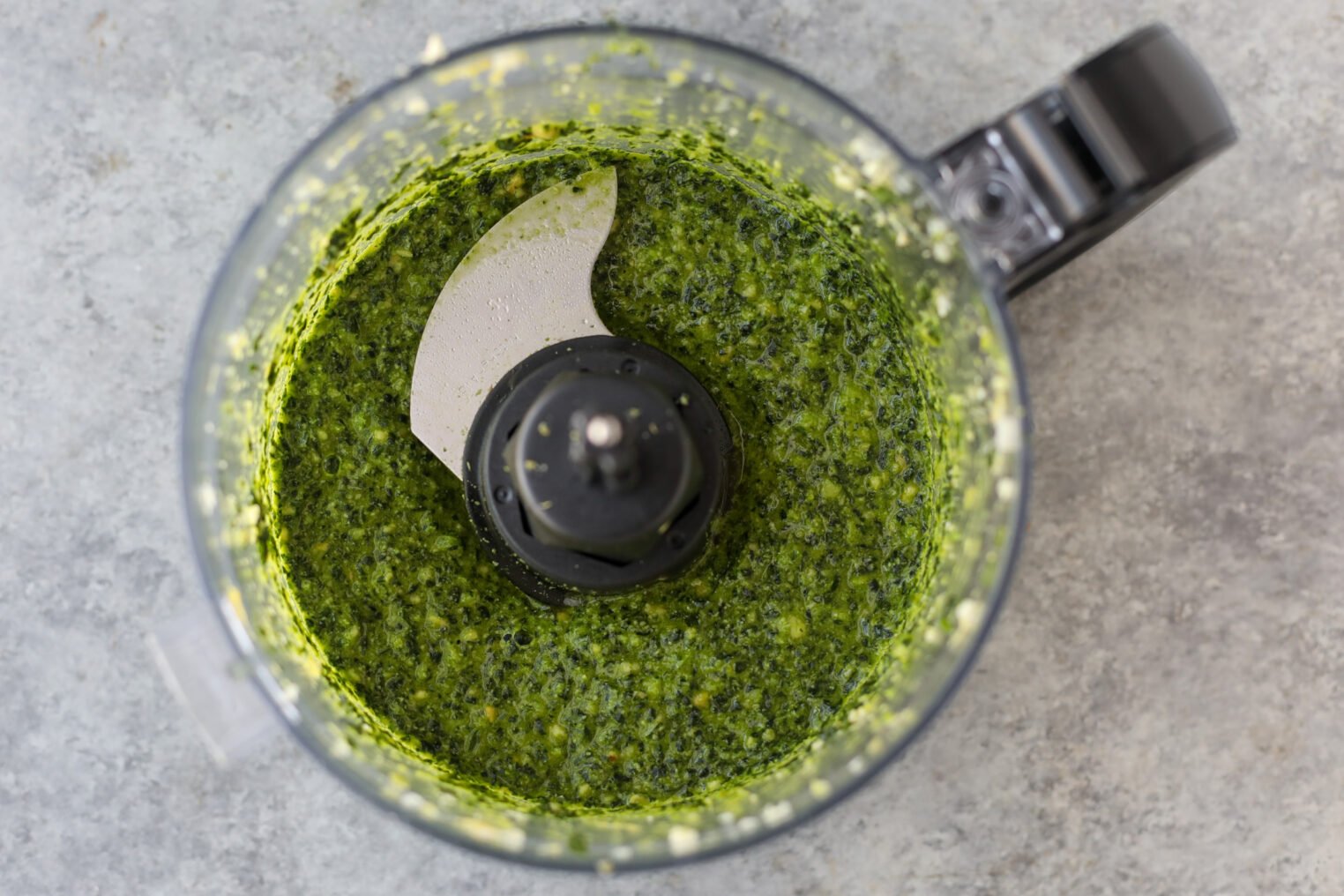 A Food Processor Bowl Filled With Freshly Blended Green Pesto Sauce, Perfect For Drizzling Over Bean Orzo Soup, Viewed From Above On A Gray Countertop.