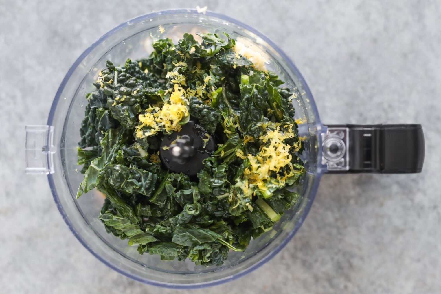 Chopped Kale And Lemon Zest Ready In A Food Processor Bowl, Perfect As A Fresh Addition To Your Bean Orzo Soup, Viewed From Above On A Gray Countertop.