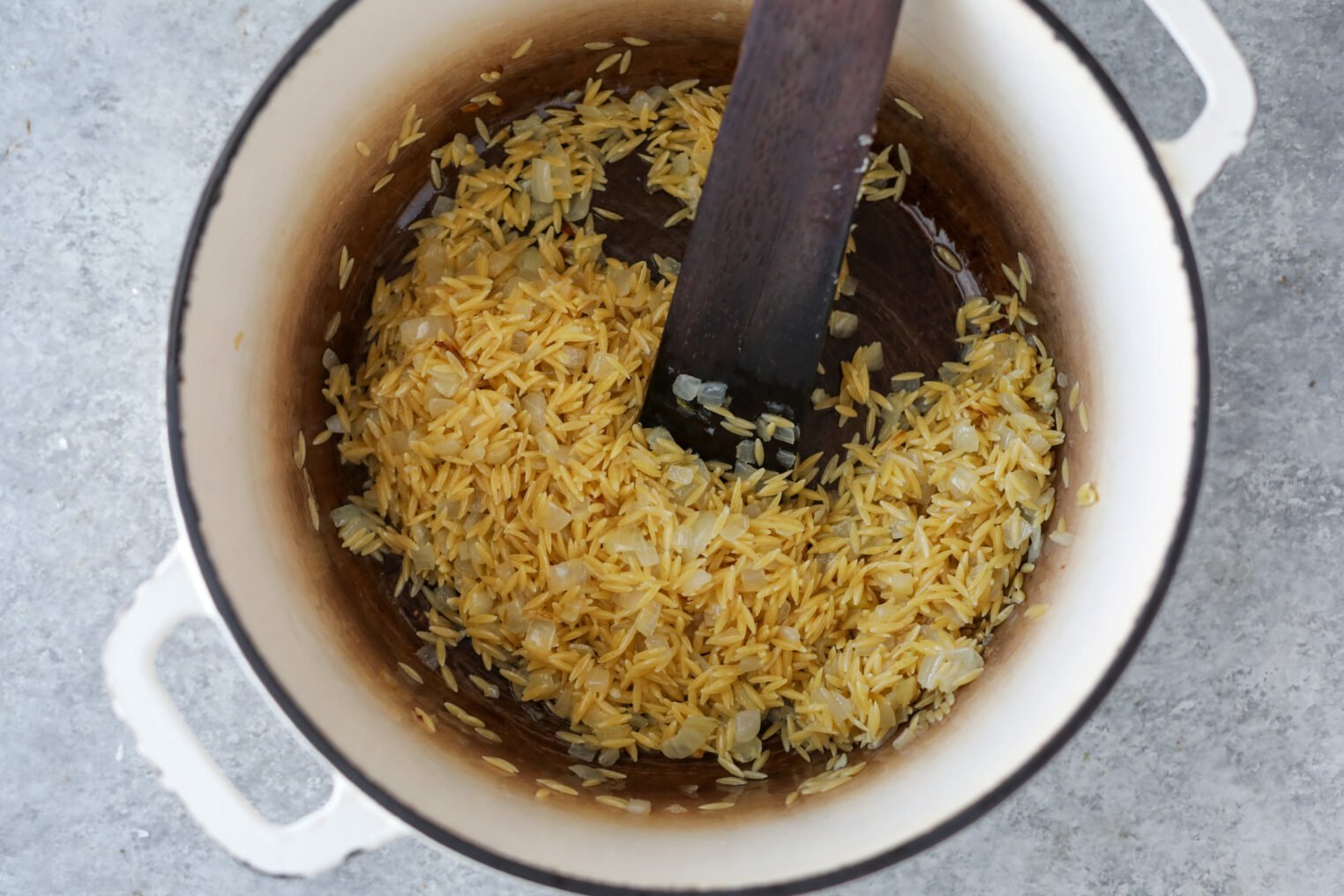 A White Pot Contains Uncooked Rice And Chopped Onions, Being Stirred With A Wooden Spatula On A Gray Countertop—Perfect Beginnings For A Hearty Bean Orzo Soup.