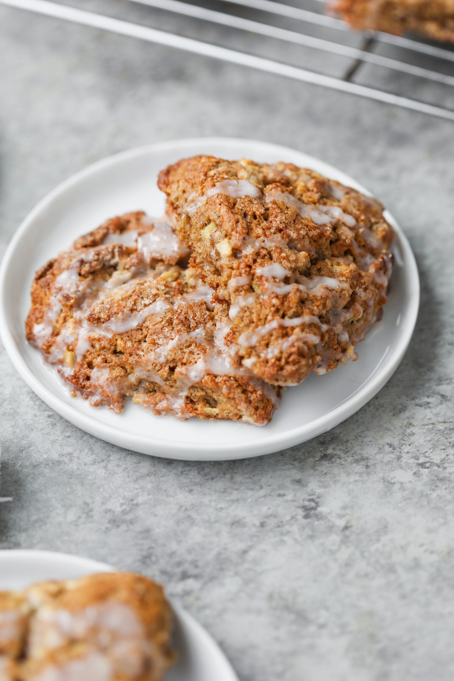 Whole Wheat Apple Scones 1 Two Golden-Brown Apple Scones With A Light Glaze Are On A Small White Plate, Set On A Gray Surface.