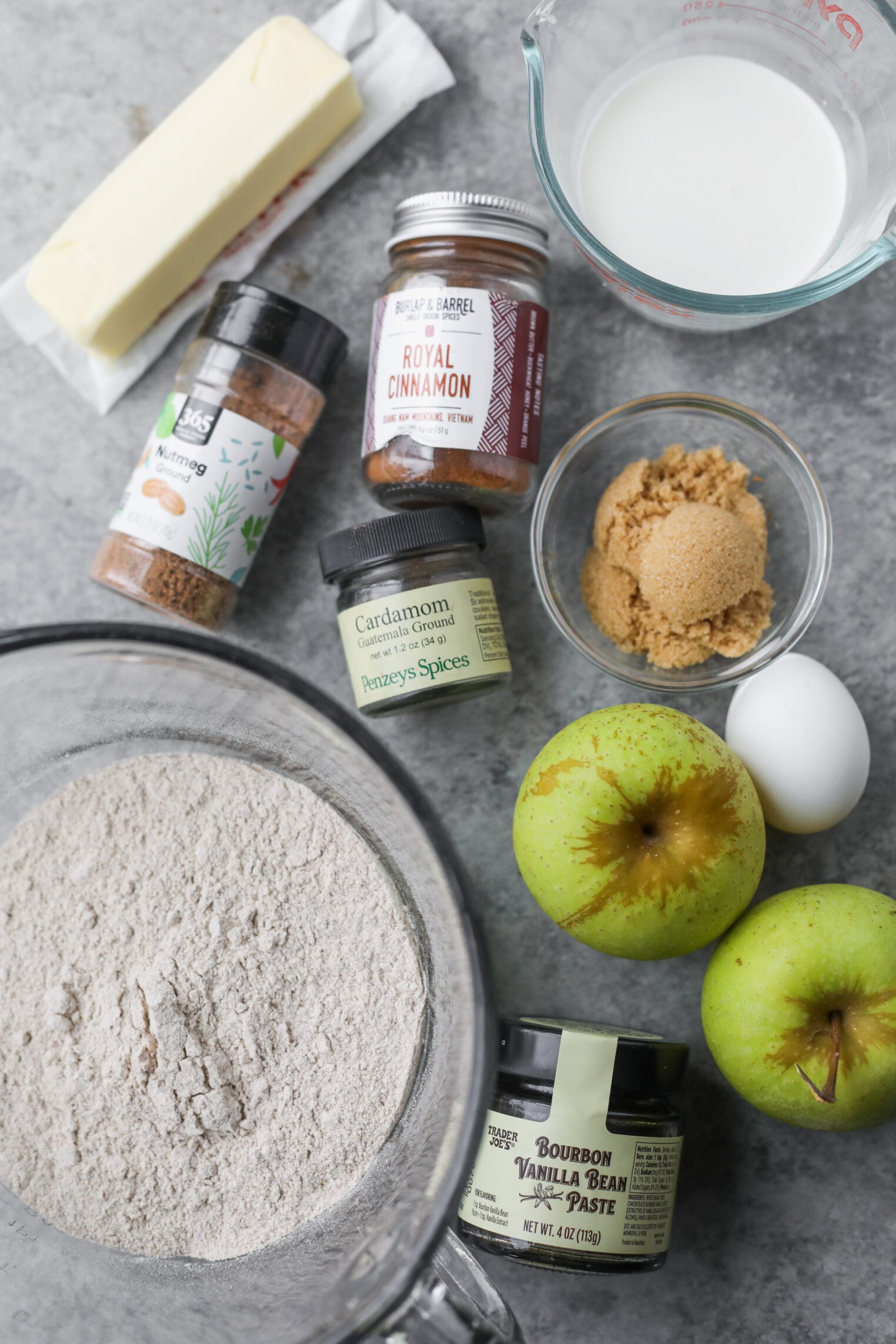 Whole Wheat Apple Scones 3 Baking Ingredients For Apple Scones On A Gray Counter: Flour In A Bowl, Two Green Apples, Eggs, Butter, Milk, Brown Sugar, Jars Of Cinnamon, Cardamom, Nutmeg, And Vanilla Bean Paste.